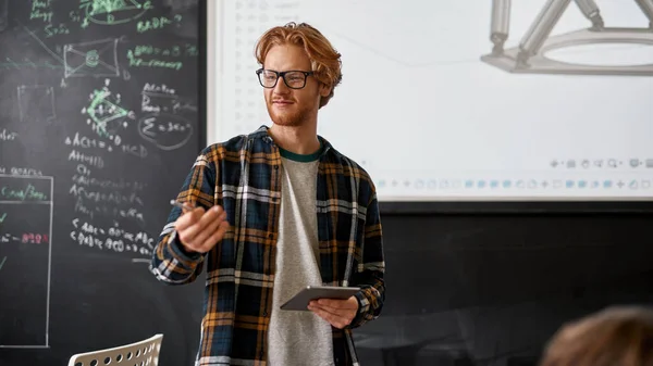 Young male caucasian teacher wearing glasses pointing on audience during teaching robotics students in classroom. Modern IT, technical and engineering education. School, college, institute