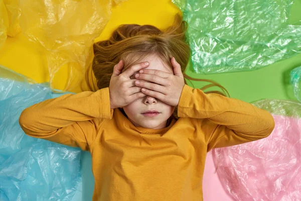 Little girl covering her eyes while lying between plastic bags on colorful background. Ecology safety and protection. Waste disposal and recycling. Environmental sustainability. Studio shoot