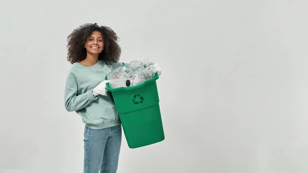 Black girl holding dustbin with plastic garbage and looking at camera. Ecology safety and protection. Waste disposal and recycling. Environmental sustainability. White background in studio. Copy space