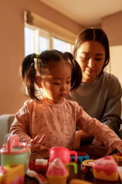 Mother looking at little daughter playing with toy figures at table at home. Domestic entertainment and leisure. Asian woman and girl spend time together. Family relationship. Parenting and motherhood