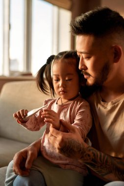 Babam evdeki kanepede dijital termometreyle kızına bakıyor. Asyalı bir adam ve küçük bir kız birlikte vakit geçiriyorlar. Sağlık ve tıbbi bakım. Aile ilişkisi. Ebeveynlik ve babalık