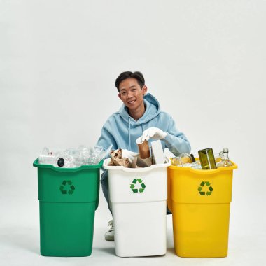 Eldiven takan Asyalı adam çöpleri cam, plastik ve kağıt kutulara diziyor ve kameraya bakıyor. Ekoloji güvenliği ve korunma. Çöp öğütücü ve geri dönüşüm. Stüdyoda beyaz geçmiş. Boşluğu kopyala