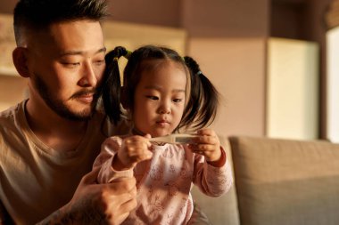 Odaklanmış baba ve kızı evdeki kanepedeki dijital termometrede ısıyı kontrol ediyor. Asyalı bir adam ve küçük bir kız. Sağlık ve tıbbi bakım. Ebeveynlik ve babalık. Modern yerli yaşam tarzı