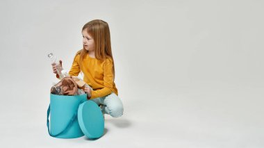 Tatmin olmamış küçük beyaz kız çöp kutusundan plastik şişeye bakıyor. Çöp ayıklama. Ekoloji güvenliği. Çöp öğütücü ve geri dönüşüm. Beyaz arka planda izole edilmiş. Stüdyo çekimi. Boşluğu kopyala