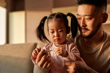 Konsantre baba ve kız evdeki kanepedeki dijital termometrede ısıyı kontrol ediyorlar. Asyalı bir adam ve küçük bir kız. Sağlık ve tıbbi bakım. Ebeveynlik ve babalık. Modern yerli yaşam tarzı