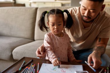 Baba ve kızı evdeki masada keçeli kalemle keçeli kalem çiziyor. Dövmeli Asyalı adam ve küçük kız birlikte vakit geçiriyorlar. Ev eğlencesi ve eğlence. Aile ilişkisi. Babalık