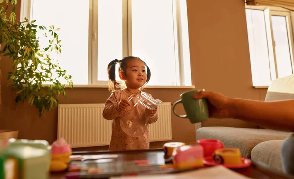 Asian little girl with glass jar looking at cropped father holding cup during they playing at home. Domestic entertainment and leisure. Family relationship. Parenting and fatherhood