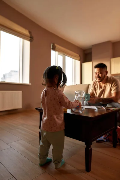 Back of girl playing with glass jar and toys with background of asian father with cup on sofa looking at girl at table at home. Domestic entertainment and leisure. Family relationship. Parenting