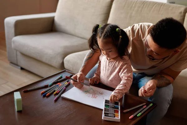 Girl taking felt-tip pen during drawing with father in copybook at table at home. Asian dad and little daughter spend time together. Domestic entertainment and leisure. Family relationship. Fatherhood
