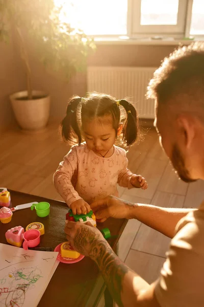 Father and asian daughter playing with toy cube with figures at table at home. Man and girl spending time together. Domestic entertainment and leisure. Family relationship. Parenting and fatherhood