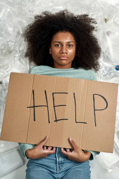 Black girl holding placard with Help word while lying between garbage. Ecology safety and protection. Waste disposal and recycling. Environmental sustainability. White background. Studio shoot