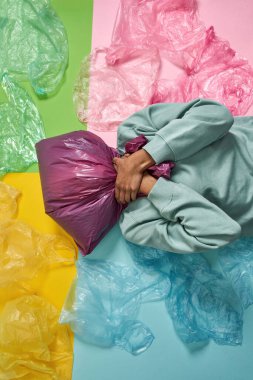 Siyah kız kafasında plastik torbayla boğulurken renkli arka planda çöplerle yatıyordu. Ekoloji güvenliği ve korunma. Çöp öğütücü ve geri dönüşüm. Çevresel sürdürülebilirlik. Stüdyo çekimi