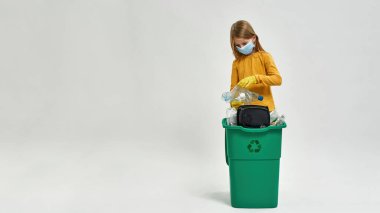 Eldiven takan ve tıbbi maske takan beyaz bir kız elinde çöp kutusundan plastik şişeler tutuyor. Çöp ayıklama. Ekoloji güvenliği. Çöp öğütücü ve geri dönüşüm. Beyaz arka plan. Stüdyo çekimi. Boşluğu kopyala