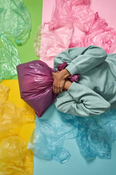 Siyah kız kafasında plastik torbayla boğulurken renkli arka planda çöplerle yatıyordu. Ekoloji güvenliği ve korunma. Çöp öğütücü ve geri dönüşüm. Çevresel sürdürülebilirlik. Stüdyo çekimi