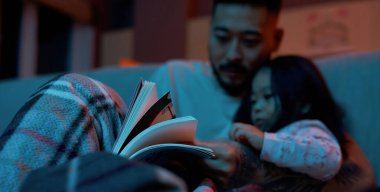 Selective focus of book in hands of blurred man reading to little daughter under blanket on sofa at home. Asian father and child spending time together. Parenting and fatherhood. Evening or night time