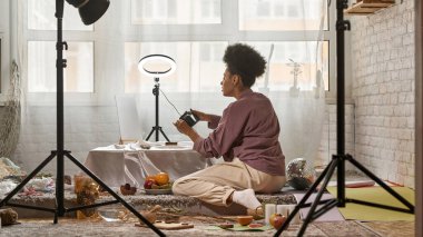 Ev stüdyosundaki dijital kamerada deniz kabuklarının fotoğrafını çeken siyahi kadın fotoğrafçının uzaktan görüntüsü. Fotostoklar, ticaret, sosyal ağlar ve reklamlar için içerik yaratan genç bir kız. Bağımsız