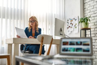 İş kadını ya da girişimci, geniş bir ofiste bilgisayarla masa başında çalışırken kağıt okuyorlar. Modern başarılı kadın yaşam tarzı. Takım elbiseli ve gözlüklü genç sarışın beyaz kadın.