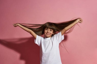 Saçlarına dokunup kameraya bakan, gülümseyen beyaz bir kız. Zoomer kuşağının dişi çocuğu. Modern gençlik yaşam tarzı kavramı. Pembe arka planda izole edilmiş. Stüdyo çekimi. Boşluğu kopyala