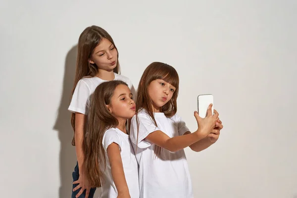 Three cute caucasian girls taking selfie on mobile phone. Friends of zoomer generation. Modern youngster lifestyle. Gadget addiction. Isolated on white background. Studio shoot. Copy space