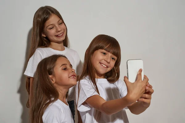 Three smiling caucasian girls taking selfie on smartphone. Friends of zoomer generation. Modern youngster lifestyle. Gadget addiction. Isolated on white background. Studio shoot. Copy space
