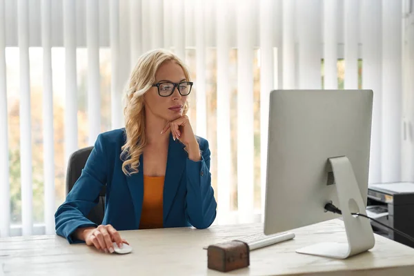 Businesswoman or company boss watching computer at desk in spacious office. Concept of modern successful female lifestyle. Young pretty focused blonde caucasian woman wearing suit and glasses
