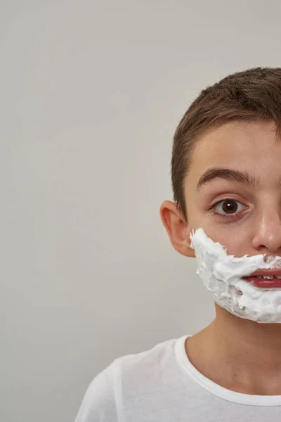 Obscure face of caucasian little boy with shaving foam on face looking at camera. Male child of zoomer generation. Modern youngster lifestyle. Skin care. White background. Studio shoot. Copy space