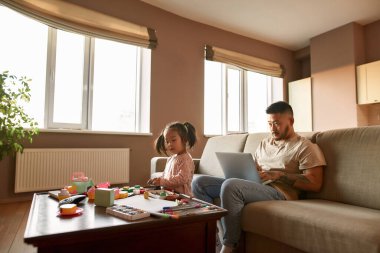 Kanepede dizüstü bilgisayarda çalışan Asyalı bir adam ve evde masada plastikle heykel yapan küçük bir kıza bakıyor. Ev eğlencesi ve eğlence. Aile ilişkisi. Serbest ve uzak çalışma