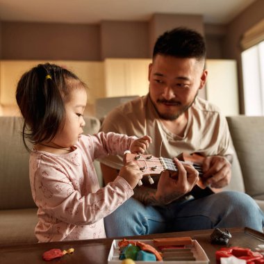 Babası evdeki kanepede müzik enstrümanında çalarken hoş bir kız ukulele kuruyor. Asyalı bir adam ve küçük bir kız. Ev eğlencesi ve eğlence. Aile ilişkisi. Babalık