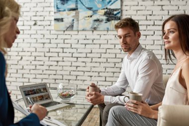 Seyahat acentesindeki genç çifte panoda birkaç tane gösteren bayan turist temsilcisi. Masasında en iyi oteller olan dizüstü bilgisayar. Müşteri hizmetleri. Tatil, turizm ve seyahat. Beyaz erkek ve kadınlar.