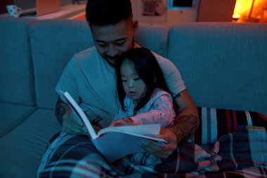 Gülümseyen baba, küçük kızıyla birlikte evde kanepenin altında kitap okuyor. Asyalı adam ve kız. Aile ilişkisi. Ebeveynlik ve babalık. Ev eğlencesi ve eğlence. Akşam ya da gece