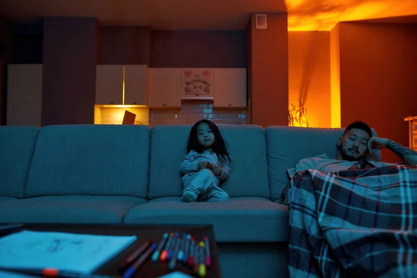 Asian little girl watching TV or movie near tired father sleeping under blanket at home. Family relationship. Parenting and fatherhood. Domestic entertainment and leisure. Evening or night time