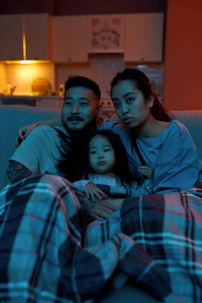 Asian family watching TV or movie under blanket on sofa at home. Mother, father and daughter spending time together. Relationship. Parenthood. Domestic entertainment and leisure. Evening or night time