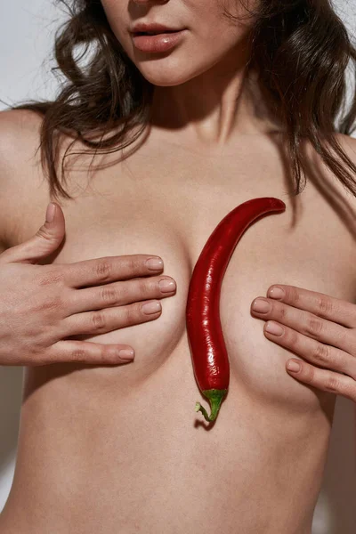 Obscure face of partial sexy nude girl holding chilli pepper between covering breast. Beautiful young slim brunette woman. Female beauty and passion. Isolated on white background. Studio shoot