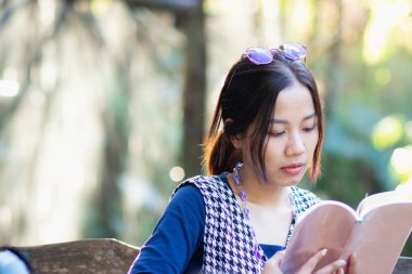 Çekici, genç Asyalı kadın doğal arka planı olan bir kitap okuyor ve rahatlatıcı bir ruh hali içinde ışıklandırıyor..