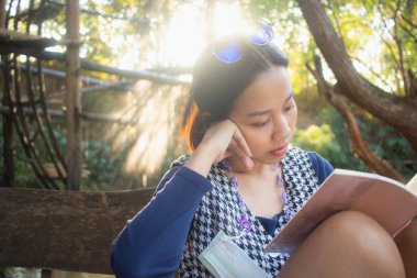 Çekici, genç Asyalı kadın doğal arka planı olan bir kitap okuyor ve rahatlatıcı bir ruh hali içinde ışıklandırıyor..