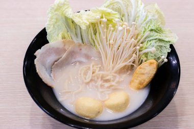 Dilimlenmiş domuz eti, napa lahanası, enoki mantarı, balık köftesi ve bıldırcın yumurtası çorbası içeren bir kase kremalı erişte çorbası. Malzemeler siyah bir kasede güzelce düzenlenmiş, rahatlatıcı bir sunum yaratıyor. 