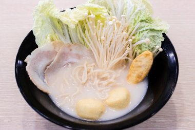 Dilimlenmiş domuz eti, napa lahanası, enoki mantarı, balık köftesi ve bıldırcın yumurtası çorbası içeren bir kase kremalı erişte çorbası. Malzemeler siyah bir kasede güzelce düzenlenmiş, rahatlatıcı bir sunum yaratıyor. 