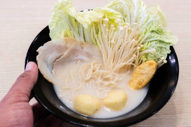 Dilimlenmiş domuz eti, napa lahanası, enoki mantarı, balık köftesi ve bıldırcın yumurtası çorbası içeren bir kase kremalı erişte çorbası. Malzemeler siyah bir kasede güzelce düzenlenmiş, rahatlatıcı bir sunum yaratıyor. 