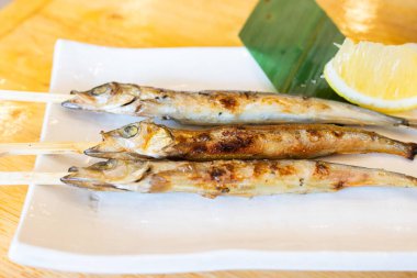 Şişler üzerinde ızgara shishamo balığı, beyaz bir seramik tabakta servis ediliyor ve arka planda bir dilim taze limon ve muz yaprağı var. Balık tamamen kömürleşmiş, altın-kahverengi bir deri sergiliyor. Geleneksel bir Japon uygulaması olarak tadını çıkarmaya hazır.