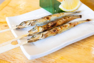 Şişler üzerinde ızgara shishamo balığı, beyaz bir seramik tabakta servis ediliyor ve arka planda bir dilim taze limon ve muz yaprağı var. Balık tamamen kömürleşmiş, altın-kahverengi bir deri sergiliyor. Geleneksel bir Japon uygulaması olarak tadını çıkarmaya hazır.