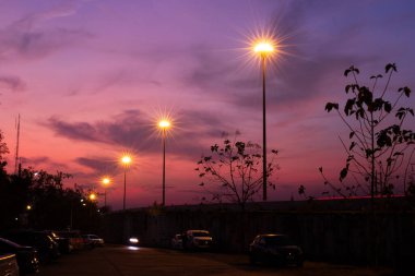 Pembe ve mor tonlarda dramatik bir günbatımı gökyüzünün altında ışıklar parlıyor. Ağaçların, arabaların ve insanların siluetleri sakin bir şehir atmosferi yaratıyor. Arka plan ve editoryal kullanım için mükemmel..