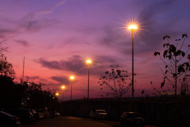 Pembe ve mor tonlarda dramatik bir günbatımı gökyüzünün altında ışıklar parlıyor. Ağaçların, arabaların ve insanların siluetleri sakin bir şehir atmosferi yaratıyor. Arka plan ve editoryal kullanım için mükemmel..