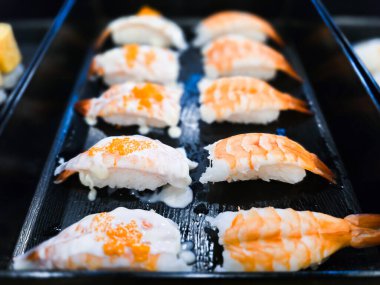 Karidesli suşi nigiri siyah bir tepside, bazıları kremalı mayonez sosu ve portakal kabuğuyla kaplı, geleneksel Japon deniz ürünleri mutfağını detaylı bir şekilde sergiliyor..