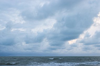 Akşamın erken saatlerinde bulutlu bir gökyüzünün altında güçlü dalgaların olduğu sakin bir sahil, kıyı boyunca uzanan çam ağaçları ve bulutların arasından süzülen yumuşak ışık.