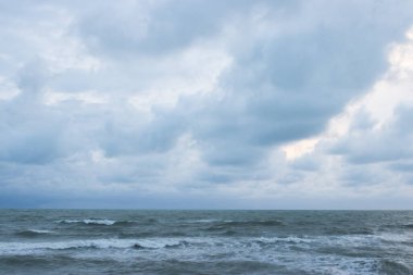 Akşamın erken saatlerinde bulutlu bir gökyüzünün altında güçlü dalgaların olduğu sakin bir sahil, kıyı boyunca uzanan çam ağaçları ve bulutların arasından süzülen yumuşak ışık.