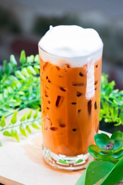 Buzlu Thai sütlü çay, kremalı ve berrak bir bardakta servis edilir, buz küpleriyle doldurulur ve arka planda yeşil yapraklarla ahşap bir yüzeye yerleştirilir..