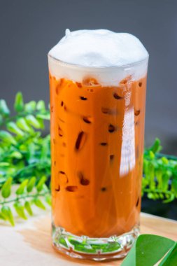 Buzlu Thai sütlü çay, kremalı ve berrak bir bardakta servis edilir, buz küpleriyle doldurulur ve arka planda yeşil yapraklarla ahşap bir yüzeye yerleştirilir..