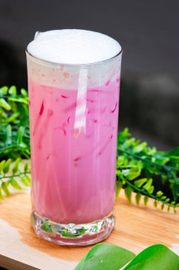 Buzlu pembe süt, kremalı, uzun bir bardakta servis edilir, buzla doldurulur ve arka planda yeşil yapraklar bulunan ahşap bir yüzeye yerleştirilir..