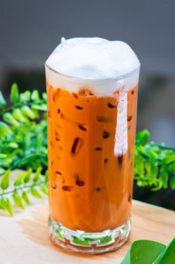 Buzlu Thai sütlü çay, kremalı ve berrak bir bardakta servis edilir, buz küpleriyle doldurulur ve arka planda yeşil yapraklarla ahşap bir yüzeye yerleştirilir..