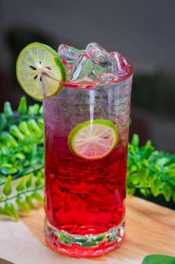 Kırmızı gazozu buzla ve limon dilimleriyle tazeleyerek berrak bir bardakta servis edilir, arka planda yeşil yapraklarla ahşap bir yüzeye yerleştirilir..